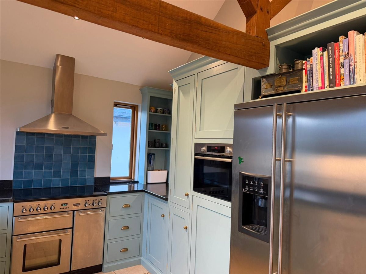 Pale kitchen cabinetry beside a range cooker and tall refrigerator beneath an exposed timber beam.