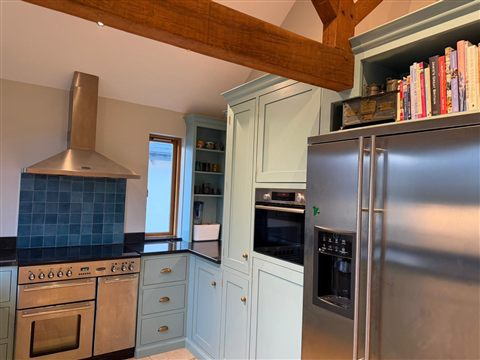 Pale kitchen cabinetry beside a range cooker and tall refrigerator beneath an exposed timber beam.