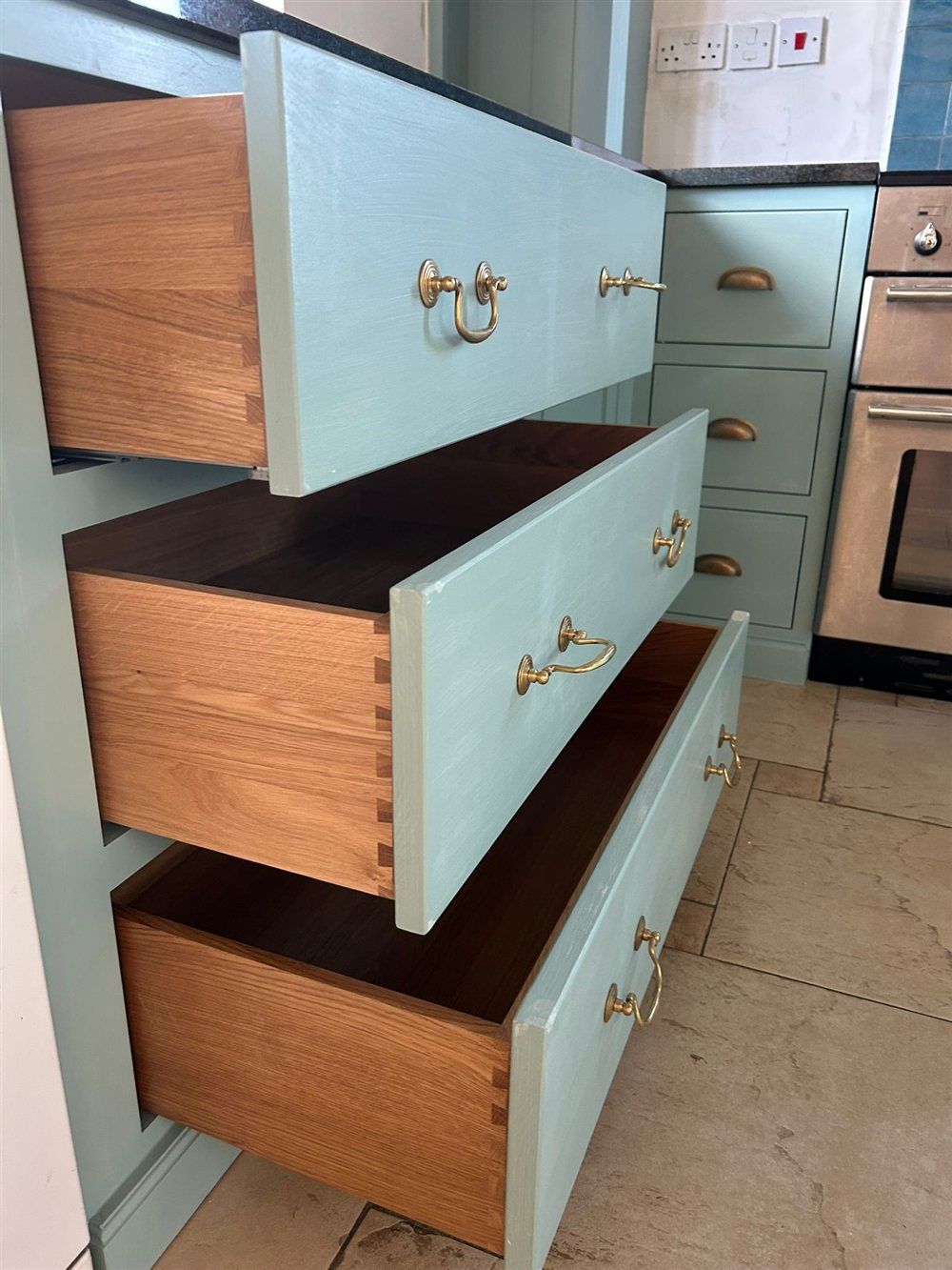 Open kitchen drawers showing solid oak dovetail joints, with brass handles on painted drawer fronts.